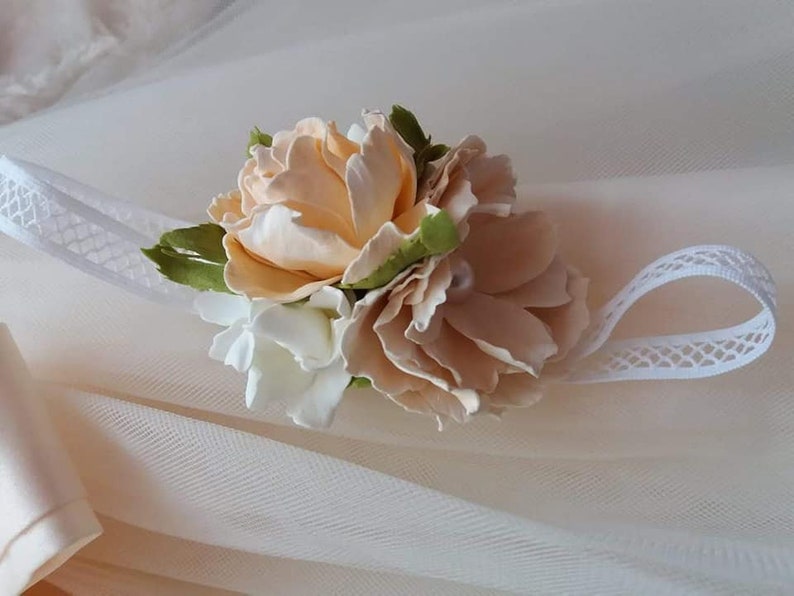 May include: A delicate white headband with a peach and white flower embellishment. The headband is made of lace and features a cluster of fabric flowers.