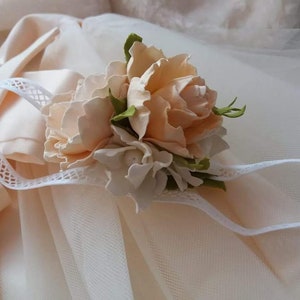 May include: A delicate white headband with a floral embellishment. The flowers are made of peach-colored fabric and have a realistic appearance. The headband is perfect for a special occasion.