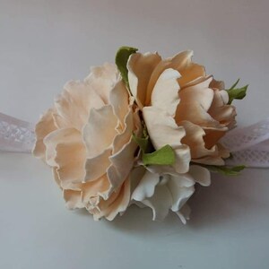 May include: A delicate floral wrist corsage featuring two peach-colored roses and a white flower, accented with green leaves. The corsage is attached to a white lace ribbon.