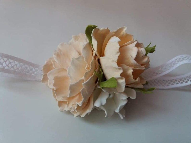 May include: A delicate floral wrist corsage featuring two peach-colored roses and a white flower, accented with green leaves. The corsage is attached to a white lace ribbon.