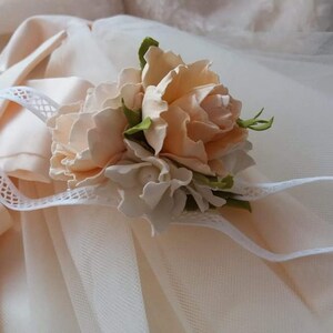 May include: A delicate white headband with a floral embellishment. The flowers are made of peach-colored fabric and have a realistic appearance. The headband is perfect for a special occasion.