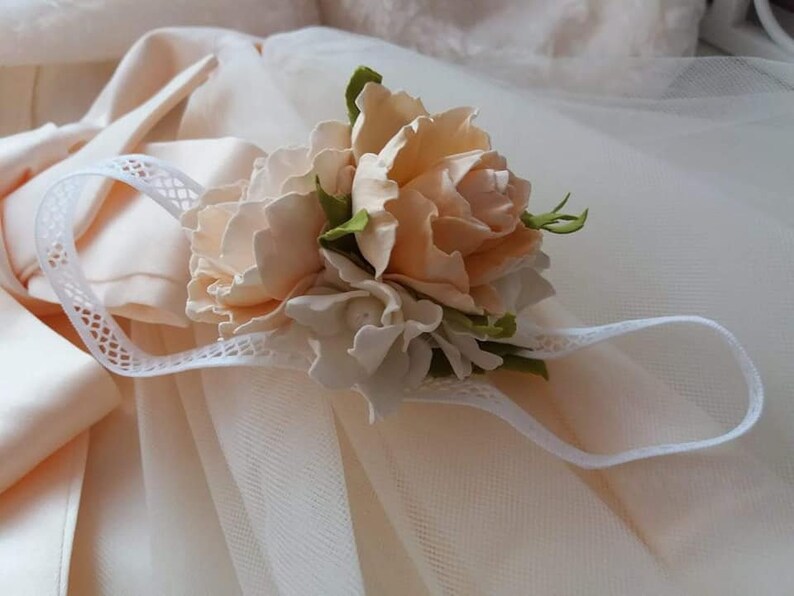 May include: A delicate white headband with a floral embellishment. The flowers are made of peach-colored fabric and have a realistic appearance. The headband is perfect for a special occasion.