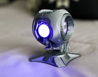 May include: A 3D printed model of a futuristic, silver-colored, spherical device with a glowing blue light emanating from the center. The device is mounted on a base with a textured surface.