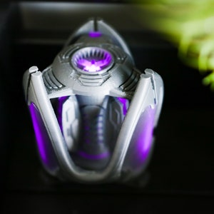 May include: A silver and purple 3D printed object with a glowing purple light inside. The object has a geometric design and appears to be a futuristic or sci-fi themed item.