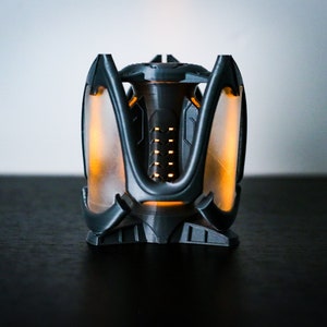 May include: A black and gray 3D printed object with a glowing orange light inside. The object has a geometric design with multiple points and a central opening.