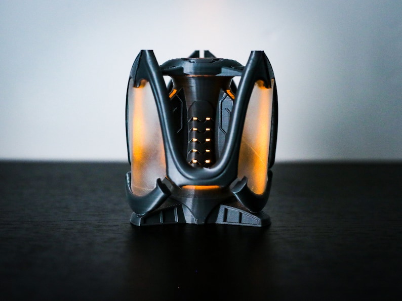 May include: A black and gray 3D printed object with a glowing orange light inside. The object has a geometric design with multiple points and a central opening.