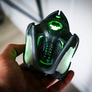 May include: A 3D printed, dark gray, futuristic-looking object with glowing green lights. The object has a complex design with intricate details and a rounded top.