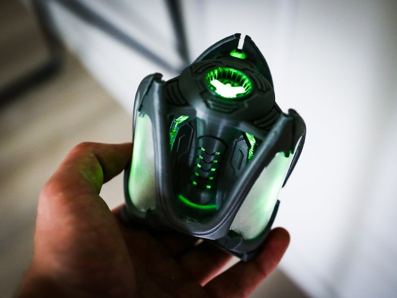 May include: A 3D printed, dark gray, futuristic-looking object with glowing green lights. The object has a complex design with intricate details and a rounded top.