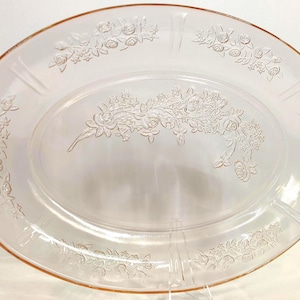 May include: Oval, light pink glass serving platter with embossed floral design. The platter features a central floral bouquet and floral patterns along the rim. The platter is a vintage piece, perfect for serving food at a dinner party.