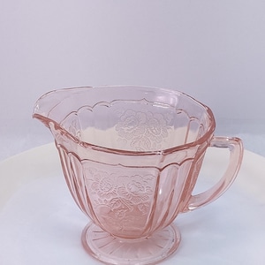 May include: A pink glass creamer with a floral pattern. The creamer has a handle and a spout. The base is round and the body is fluted. The creamer is made of glass and is a light pink color.