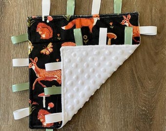 May include: A black and white baby tag blanket with a white minky backing. The front features a woodland animal print with foxes, deer, squirrels, mushrooms, and butterflies. The blanket has multiple satin ribbon tags attached to the edges.