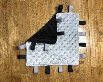 May include: A grey and black baby tag blanket with a soft, textured surface. The blanket has a square shape with a black, plush corner and a grey, dimpled fabric. Black and grey ribbons are attached around the edges.