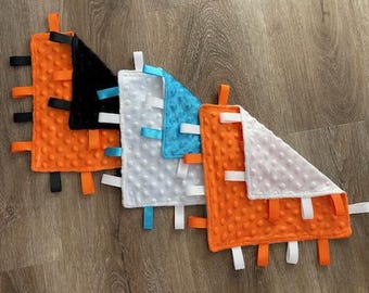 May include: Five different baby blankets in a range of colours and textures. The blankets are made from soft, plush fabric and have satin ribbons attached to the corners. The blankets are arranged in a row, with the orange blanket at the bottom and the black blanket at the top.