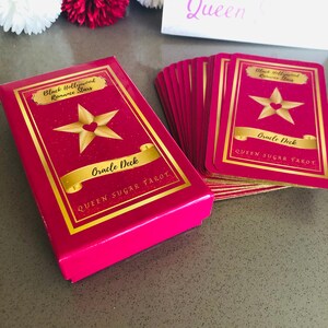 May include: A pink Oracle Deck box and cards with gold lettering and a star design. The box and cards read "Black Hollywood Romance Stars Oracle Deck QUEEN SUGAR TAROT". The cards are fanned out next to the box.