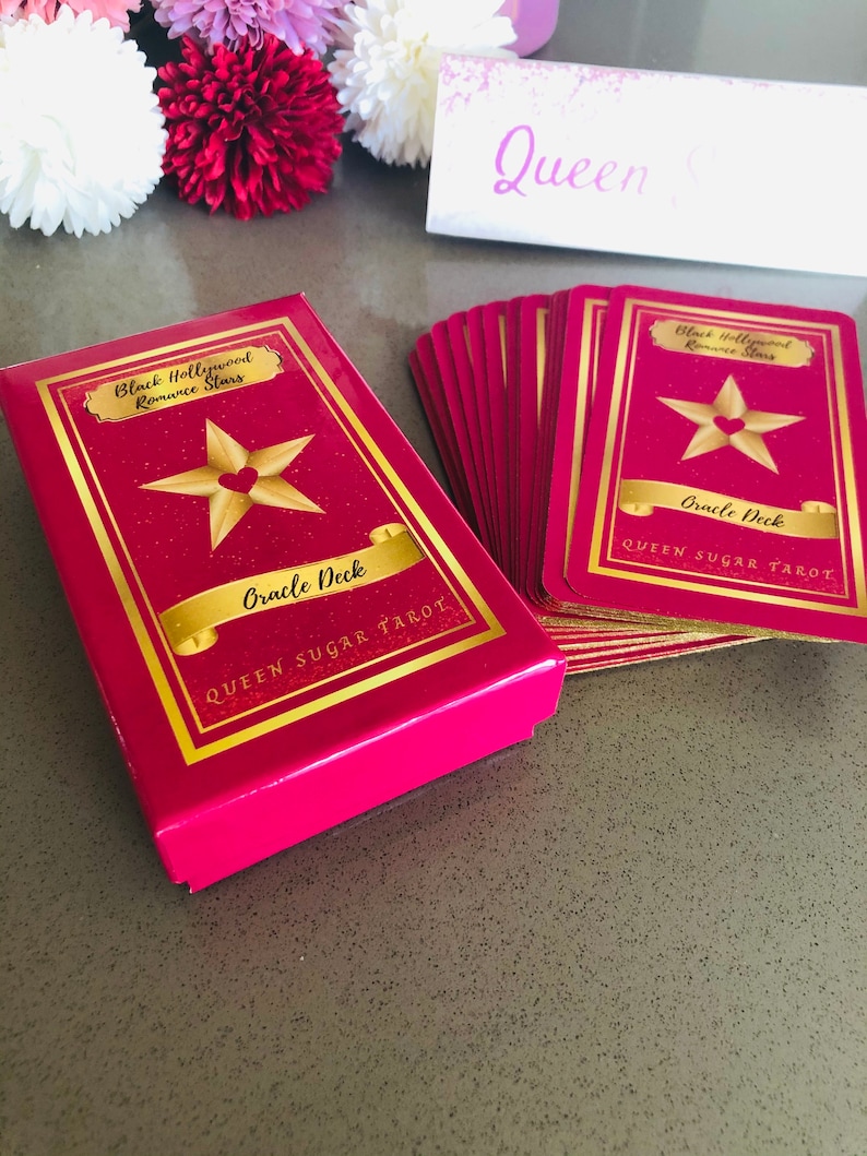 May include: A pink Oracle Deck box and cards with gold lettering and a star design. The box and cards read "Black Hollywood Romance Stars Oracle Deck QUEEN SUGAR TAROT". The cards are fanned out next to the box.