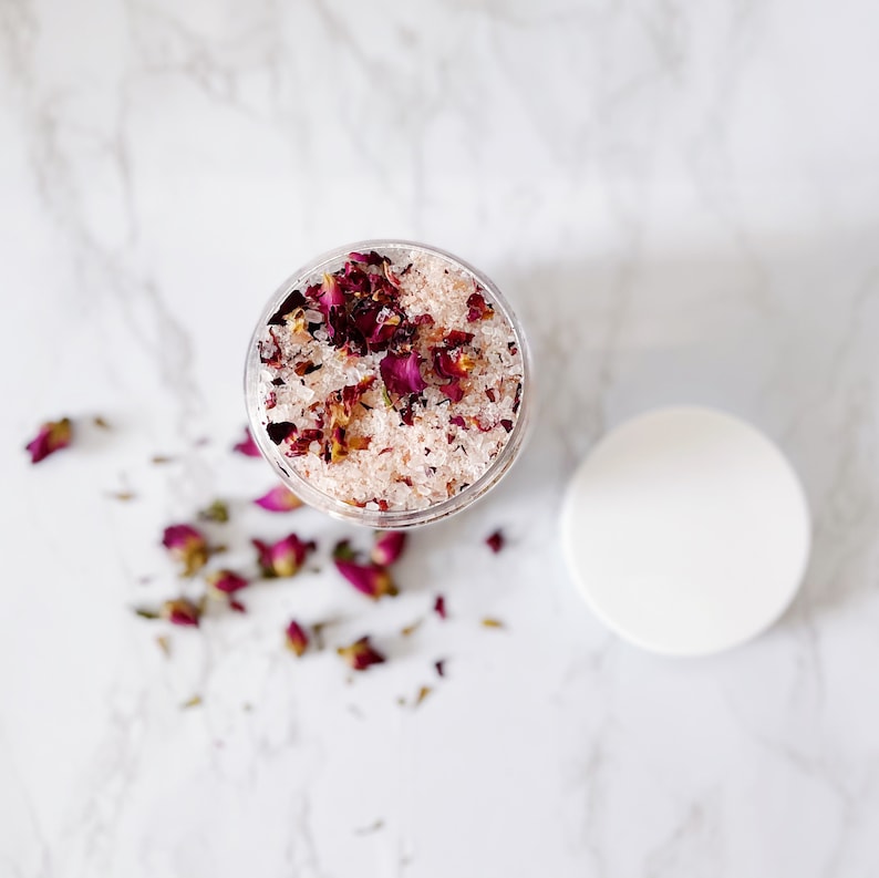 May include: A clear glass jar filled with pink bath salts and dried rose petals. The jar is on a white marble surface.