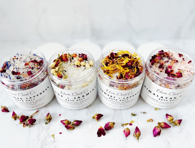 May include: Four glass jars filled with bath salts and dried flowers. The jars are labeled with the names of the scents: Eucalyptus Lavender, Rose Jasmine, Rosemary Lavender, and Himalayan Pink.