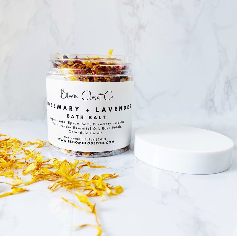 May include: A clear glass jar filled with bath salts, rose petals, and calendula petals. The jar has a white label with the text "Bloom Closet Co. Rosemary + Lavender Bath Salt" and the ingredients: "Epsom Salt, Rosemary Essential Oil, Lavender Essential Oil, Rose Petals, Calendula Petals". The jar is sitting on a white marble surface with yellow flower petals scattered around it. The net weight is 8.5 oz (241 g).