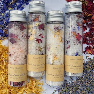 May include: Four small glass bottles filled with bath salts and dried flowers. The bottles are labeled with the text "Bloom Closet Co" and the names of the different bath salts: "Himalayan Rose Bath Salt", "Rose & Jasmine Bath Salt", "Rosemary & Lavender Bath Salt", and "Luxurious Lavender Bath Salt". Each bottle contains 1.5 oz (42 g) of bath salts.