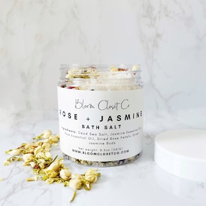 May include: A clear glass jar filled with bath salts, dried rose petals, and jasmine flowers. The label reads "Bloom Closet Co Rose + Jasmine Bath Salt" and lists the ingredients: Dead Sea Salt, Jasmine Essential Oil, Dried Rose Petals, Dried Jasmine Buds. The net weight is 8.5 oz (241g). The website address is www.bloomclosetco.com.