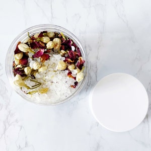 May include: A clear glass jar filled with white bath salts, dried rose petals, and white jasmine flowers. The jar has a white lid.