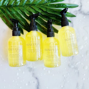 Pu&ograve; includere: Quattro bottiglie trasparenti di olio per il corpo Honey Glow con erogatori a pompa neri. Le bottiglie sono su una superficie di marmo bianco con una foglia verde sullo sfondo. Le bottiglie contengono un liquido giallo e l'etichetta dice "Honey Glow Body Oil".