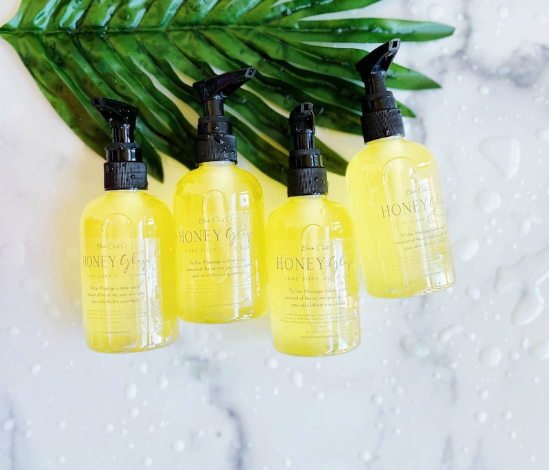 Pu&ograve; includere: Quattro bottiglie trasparenti di olio per il corpo Honey Glow con erogatori a pompa neri. Le bottiglie sono su una superficie di marmo bianco con una foglia verde sullo sfondo. Le bottiglie contengono un liquido giallo e l'etichetta dice "Honey Glow Body Oil".