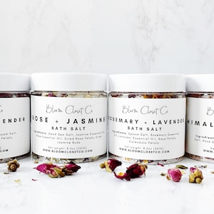 May include: Four glass jars of bath salts with white lids and labels. The labels are white with black text and list the scents of the bath salts: Furious Lavender, Rose + Jasmine, Rosemary + Lavender, and Himalayan Rose. The jars are sitting on a white surface with dried rose petals scattered around them.