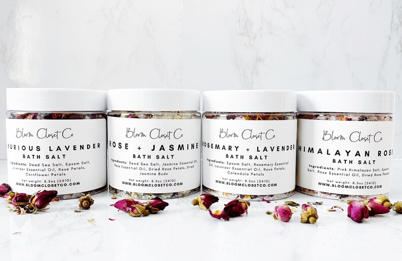 May include: Four glass jars of bath salts with white lids and labels. The labels are white with black text and list the scents of the bath salts: Furious Lavender, Rose + Jasmine, Rosemary + Lavender, and Himalayan Rose. The jars are sitting on a white surface with dried rose petals scattered around them.