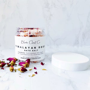 May include: A glass jar filled with pink Himalayan bath salts, dried rose petals, and rose essential oil. The jar has a white label with the text "Bloom Closet Co. Himalayan Rose Bath Salt" and the ingredients listed. The jar is sitting on a white marble surface with dried rose petals scattered around it.