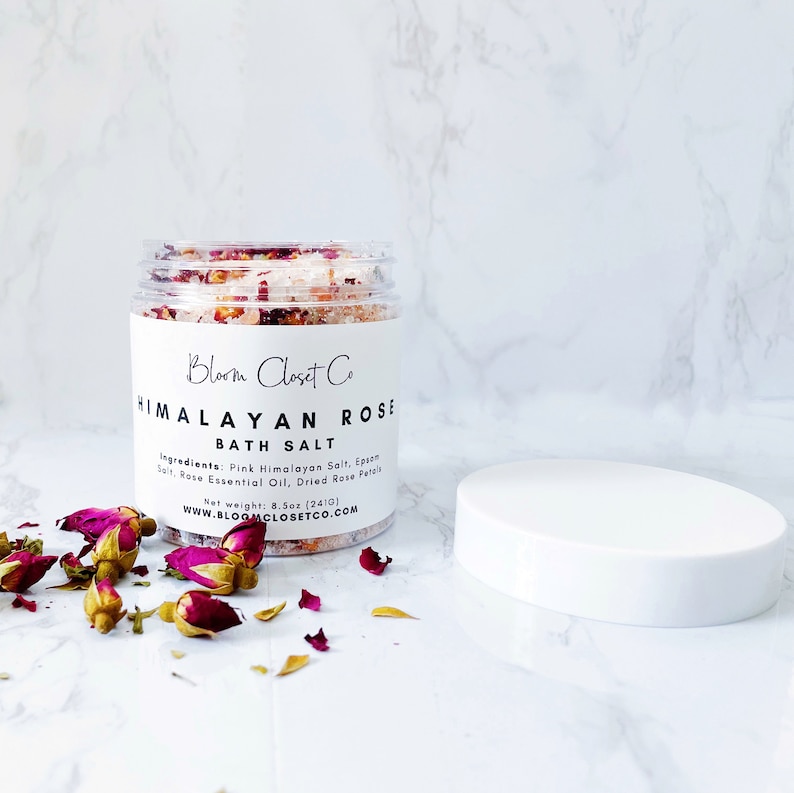 May include: A glass jar filled with pink Himalayan bath salts, dried rose petals, and rose essential oil. The jar has a white label with the text "Bloom Closet Co. Himalayan Rose Bath Salt" and the ingredients listed. The jar is sitting on a white marble surface with dried rose petals scattered around it.