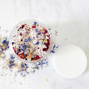 May include: A clear glass jar filled with white bath salts, pink rose petals, and blue flower petals. The jar is on a white marble surface.