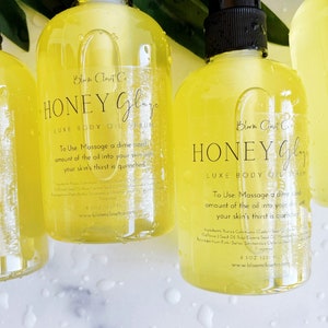 Pu&ograve; includere: Tre bottiglie di vetro ambrato di siero olio per il corpo Honey Glaze Luxe. Le bottiglie hanno etichette nere con testo bianco che recita "Honey Glaze Luxe Body Oil Serum" e istruzioni per l'uso. Le bottiglie sono su una superficie bianca con gocce d'acqua.