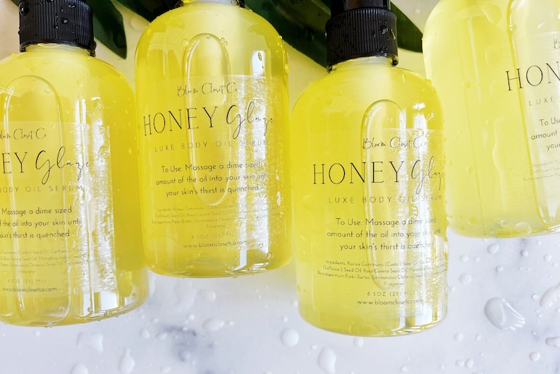 Pu&ograve; includere: Tre bottiglie di vetro ambrato di siero olio per il corpo Honey Glaze Luxe. Le bottiglie hanno etichette nere con testo bianco che recita "Honey Glaze Luxe Body Oil Serum" e istruzioni per l'uso. Le bottiglie sono su una superficie bianca con gocce d'acqua.