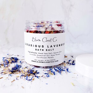 May include: A clear glass jar filled with white bath salts, lavender, rose, and cornflower petals. The jar has a white label with the text "Bloom Closet Co. Luxurious Lavender Bath Salt" and the ingredients and net weight. The label also includes the website address "www.bloomclosetco.com".