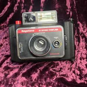 May include: Black Keystone 60 Second Everflash instant camera with a red button and a built-in flash. The camera has a silver lens ring and a dial for adjusting the lighting. The text "Keystone" and "60 Second Everflash" are visible.