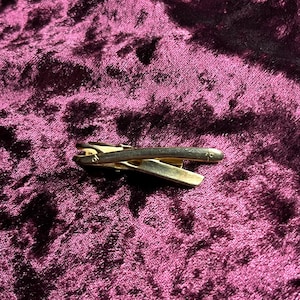 Puede incluir: Un clip de corbata dorado con un diseño sencillo y elegante. El clip tiene forma rectangular con la parte superior redondeada y un pequeño elemento decorativo en un extremo. El fondo es de terciopelo burdeos oscuro.