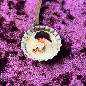May include: A circular pendant with a vintage image of a woman and a butterfly, set in a silver bottle cap. It hangs from a brown leather cord. The background is a deep purple velvet fabric.
