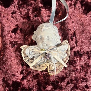 Op de afbeelding: Een lichtblauw cherubijnornament met een kanten kraag en strik. De cherubijn heeft gedetailleerde gelaatstrekken en krullend haar. Een lichtblauw lint is bevestigd om op te hangen. Het ornament staat tegen een bordeauxrode fluwelen achtergrond.
