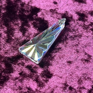 May include: A clear, prism-shaped crystal pendant with a silver-colored metal clasp. The crystal has a fan-like design at the base and reflects multiple colors. The pendant is displayed on a deep purple velvet surface.