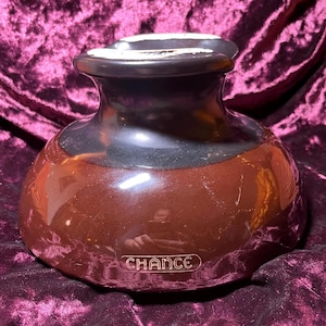 Puede incluir: Un jarrón de cerámica marrón con acabado brillante. El jarrón tiene una base ancha y redondeada que se estrecha hacia un cuello más estrecho y un borde acampanado. La palabra "CHANCE" está grabada en relieve en la parte delantera. El jarrón es probablemente vintage.