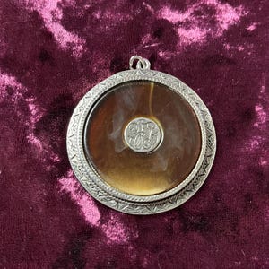 May include: A round, silver-toned pendant with a brown, circular center. The center has a small, silver-toned emblem in the middle. The pendant has a decorative, patterned border and a loop at the top for a chain.