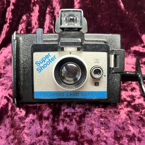 Puede incluir: Una cámara Polaroid Super Shooter Land vintage. La cámara es negra con una cara plateada y un accesorio de flash. Las palabras "Super Shooter" y "Polaroid Land Camera" están impresas en la parte delantera.
