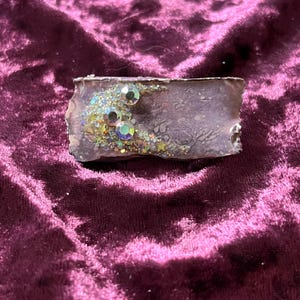 May include: A rectangular, lavender-coloured bar with a textured surface. The bar is adorned with iridescent glitter and three large, clear, round rhinestones. The edges of the bar have a rough, uneven appearance. The bar is set against a deep purple velvet background.