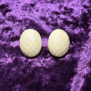 May include: A pair of oval-shaped earrings in a cream color, featuring a faceted diamond-cut pattern across the surface. The earrings have a smooth, polished finish and a rounded, convex profile that reflects light across the geometric facets.
