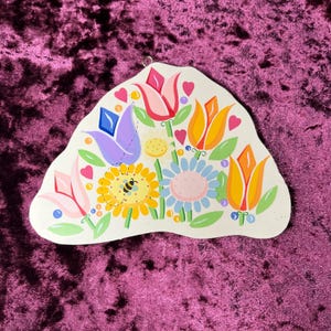 May include: A hand-painted, off-white wooden wall hanging featuring a colorful floral design. The artwork includes tulips, daisies, and hearts in shades of pink, yellow, orange, blue, and purple. A small bee is painted on a yellow flower.