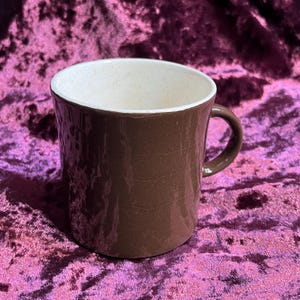 May include: A brown ceramic mug with a textured exterior and a white speckled interior. The mug has a curved handle and is set against a purple background. This mug is suitable for coffee, tea, or other beverages.