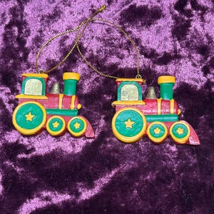 Puede incluir: Dos adornos de tren con un esquema de color rojo, verde y amarillo. Cada locomotora tiene una ventana de color dorado, una pequeña campana en la parte superior y tres ruedas decoradas con estrellas amarillas. Un hilo dorado fino cuelga de cada adorno.