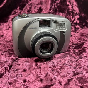 Könnte beinhalten: Silberne Argus Digitalkamera mit Zoomobjektiv. Die Kamera hat einen schwarzen Griff und einen Objektivring. Das Objektiv ist mit "Aspherical Zoom Lens" und "16mm-55mm" beschriftet. Die Kamera liegt auf einer weinroten Oberfläche.