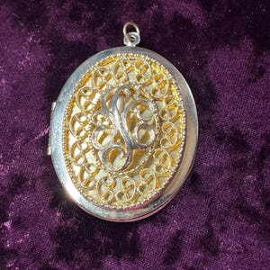 May include: Oval-shaped locket with a silver frame and a gold filigree design. The center features a silver letter "J". The locket has a small loop at the top for a chain. The background is a deep purple velvet.